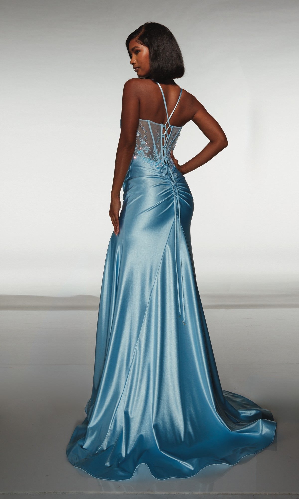 A woman stands with one hand on her hip, modeling the Alyce 62059 long formal dress—a floor-length light blue satin gown with an embellished corset bodice and lace-up back—against a plain background.